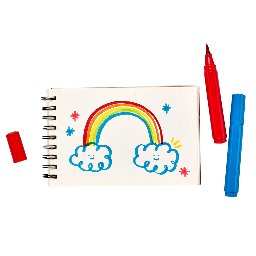 Een tekening van een regenboog met lachende wolken in een schetsboek, met twee ooly viltstiften ernaast.