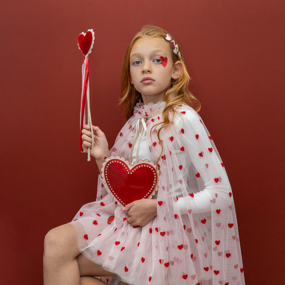 Meisje met rood haar houdt de toverstaf en een rood hart vast, zittend voor een rode achtergrond.