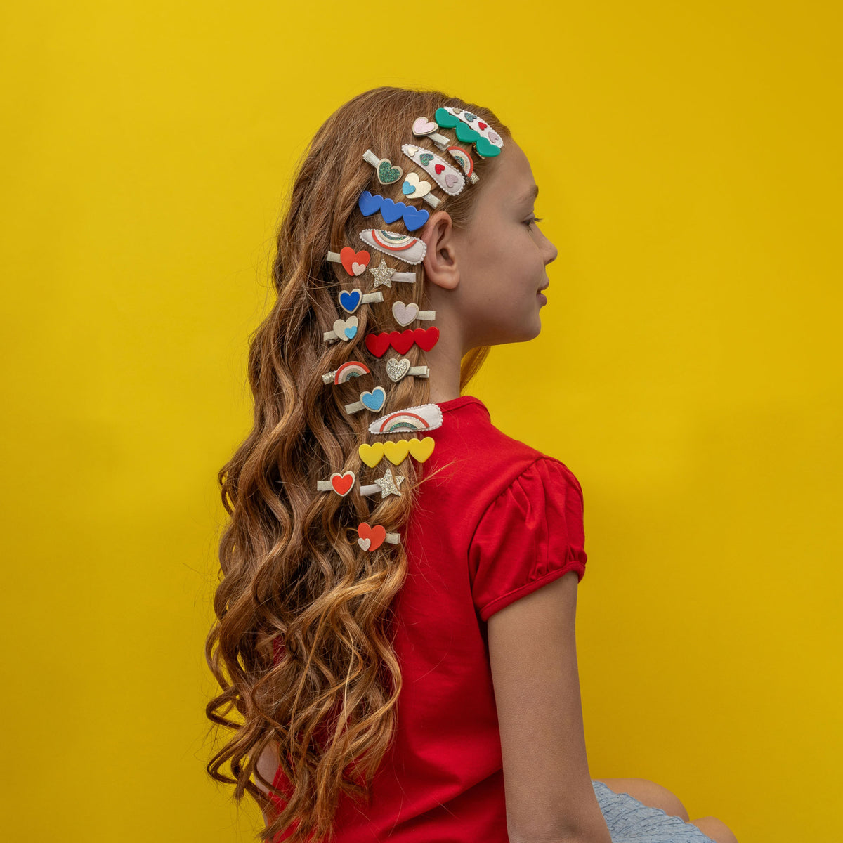 Een meisje met lang haar draagt diverse haarclipjes, waaronder de mimi & lula regenboog en harten haarclipjes, tegen een gele achtergrond.
