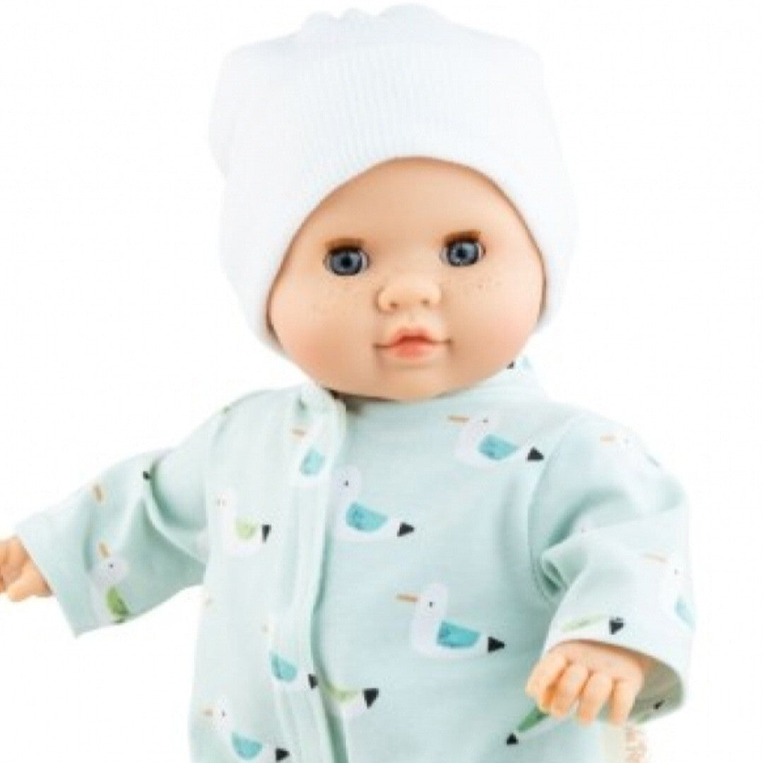 Detail van de Paola Reina babypop Manus Martin met blauwe ogen, een lichtblauwe jumpsuit met vogelpatroon en een witte muts.