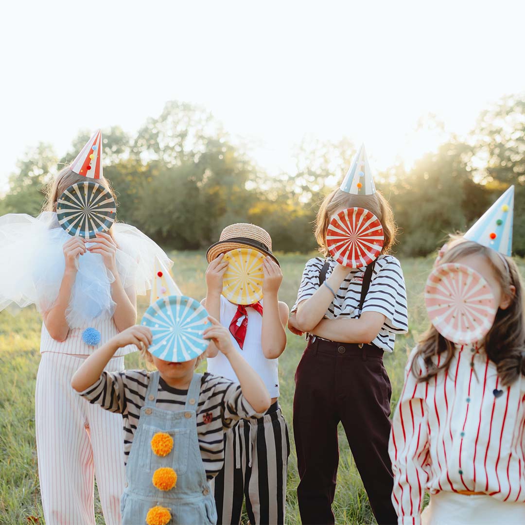 Vijf kinderen dragen PartyDeco feesthoedjes en houden ronde bordjes voor hun gezicht in een grasveld.
