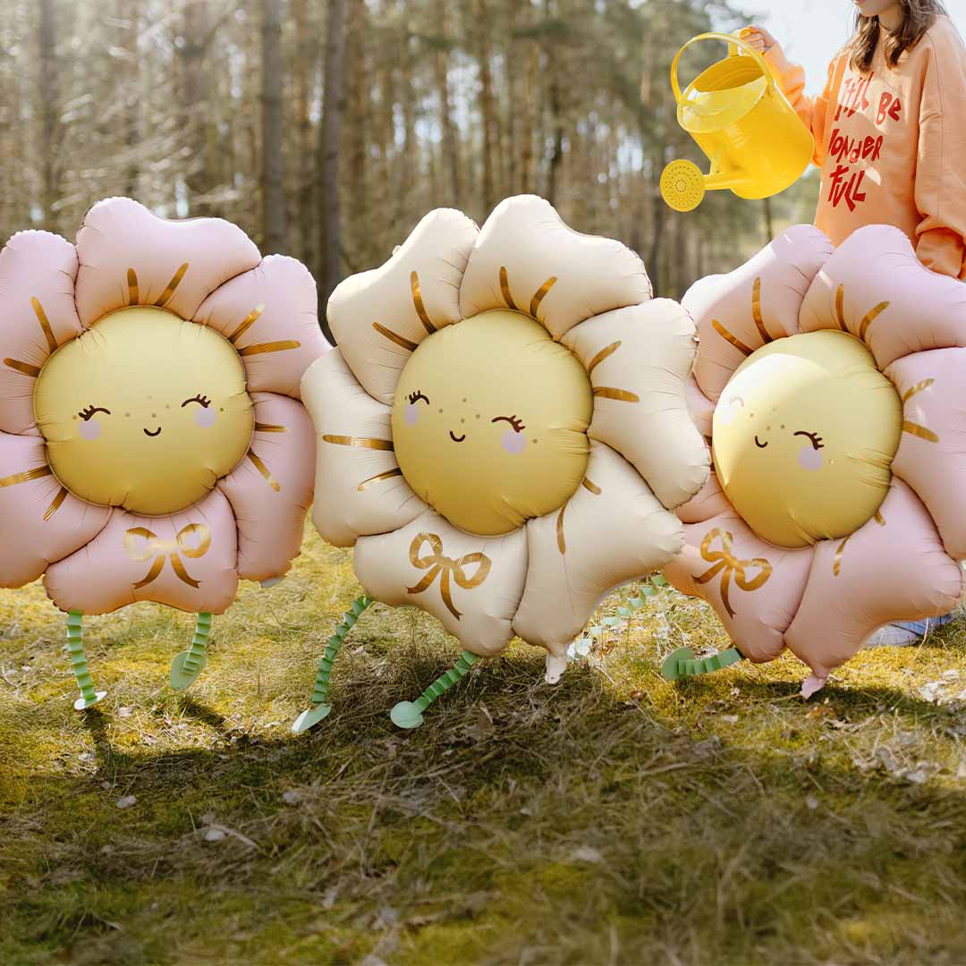 Drie partydeco folieballonnen in de vorm van bloemen, twee roze en één beige, staan in een bos.