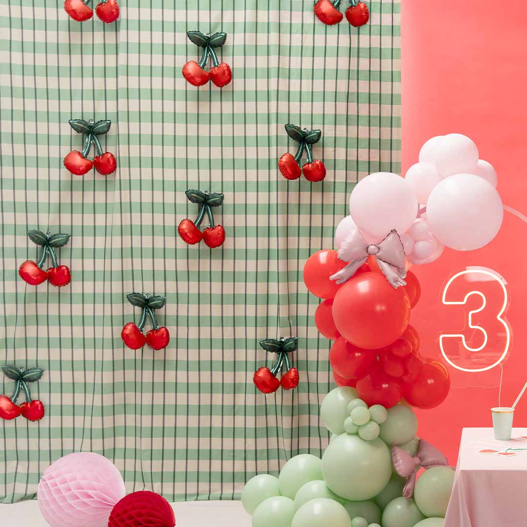 Partydeco kersen folieballonnen als decoratie op een feestje met een groene geruite wand en andere ballonnen.