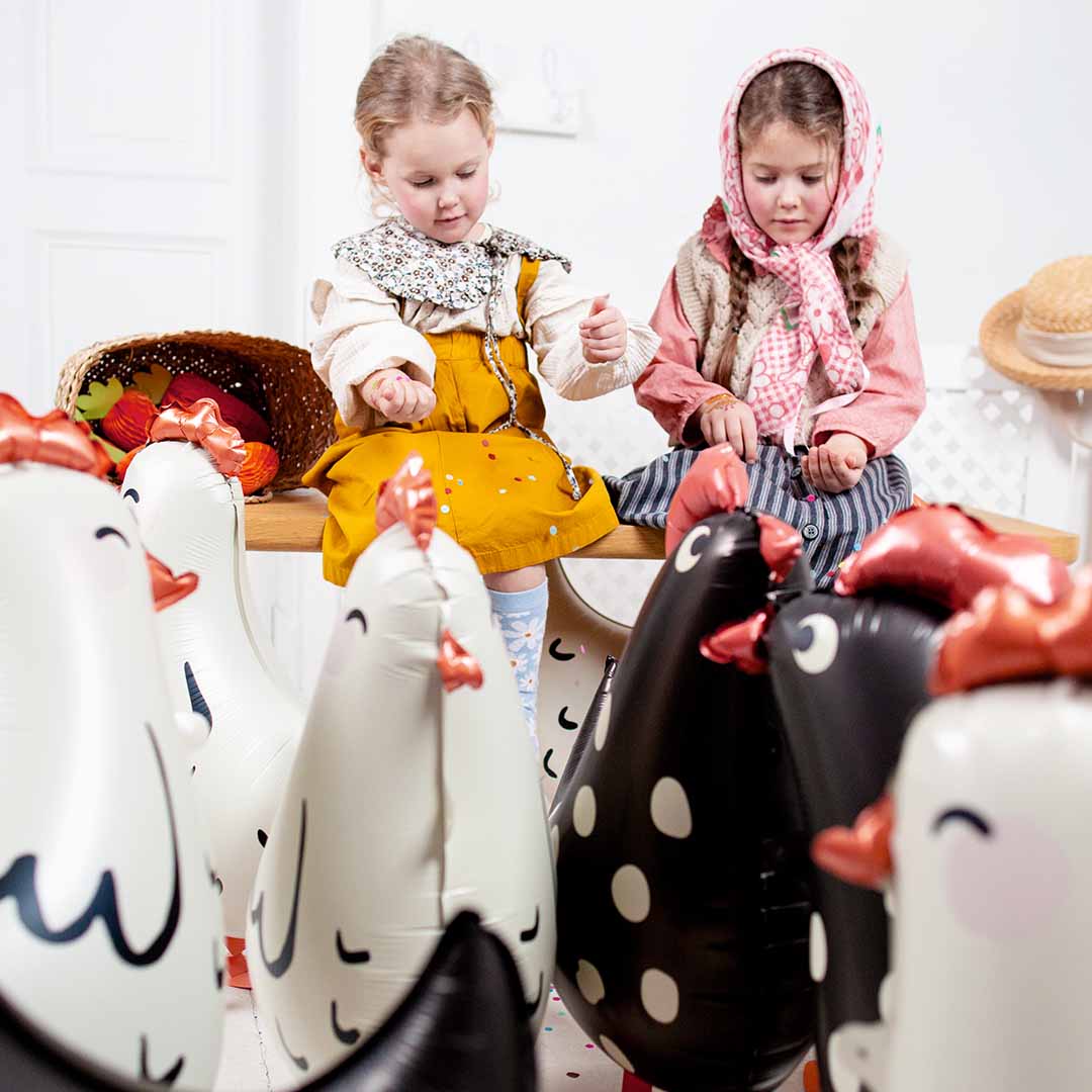 Twee meisjes kijken naar Partydeco folieballonnen in de vorm van witte en zwarte kippen die op de grond staan.