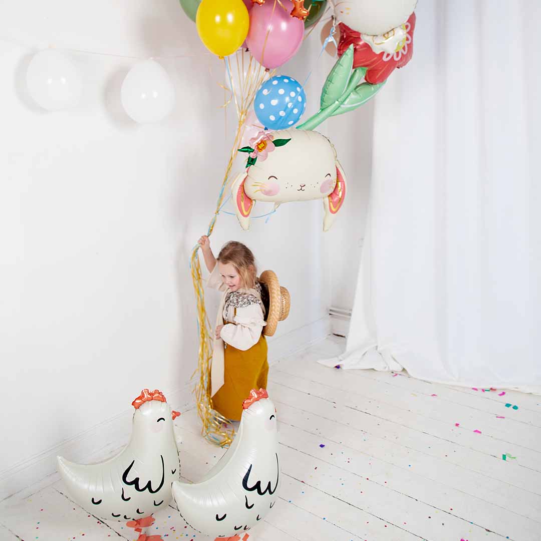 Een meisje houdt een tros ballonnen vast, met twee Partydeco folieballonnen in de vorm van witte kippen op de grond.