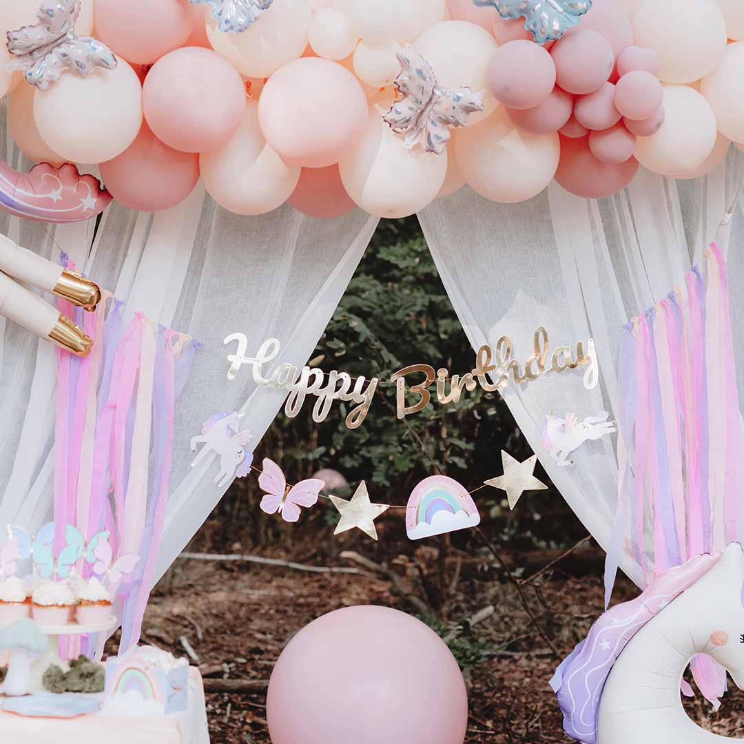 Partydeco feestslinger met 'Happy Birthday' tekst en eenhoornfiguren, hangend in een feestelijke setting met roze ballonnen.