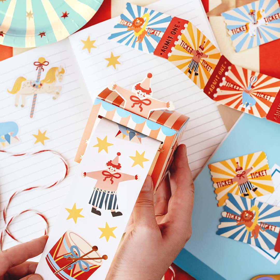 Handen trekken een rol partydeco circus stickers uit een gestreepte doos, met losse tickets en feestartikelen op de achtergrond.