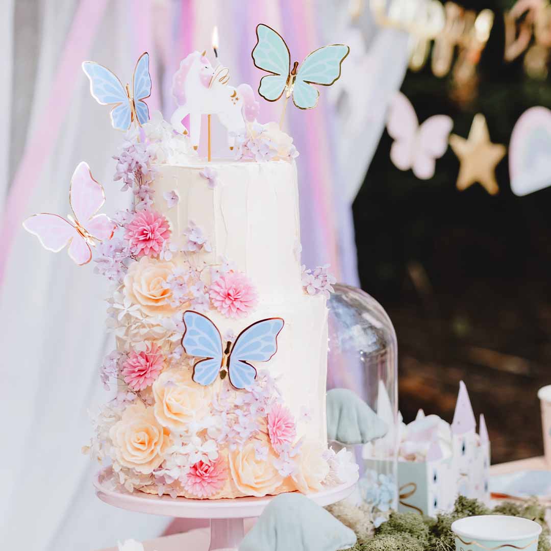 Een feesttaart met partydeco vlinder- en eenhoorn taarttoppers en bloemen op een buitentafel.