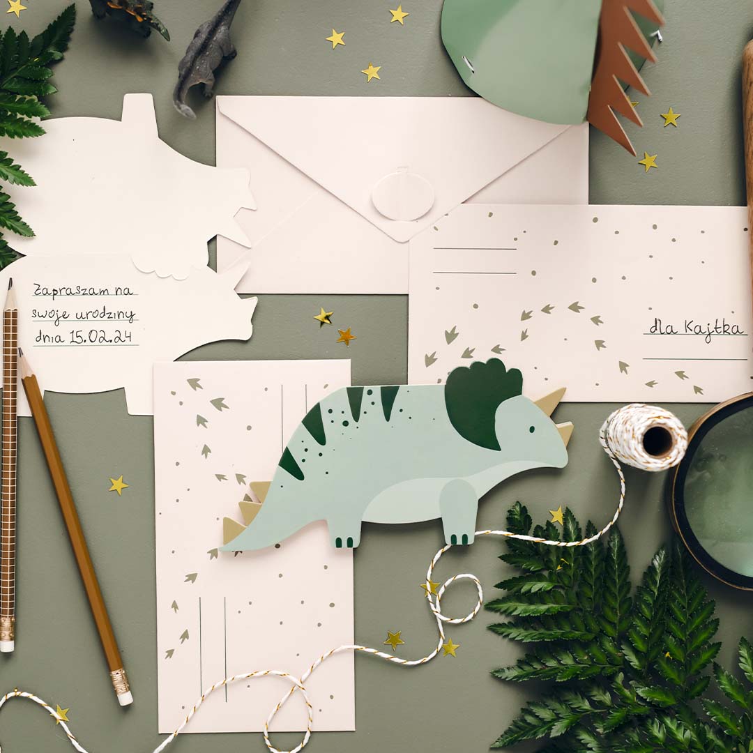 Partydeco triceratops verjaardagsuitnodigingen en enveloppen, gedemonstreerd met schrijfwaren en decoratie op een groene ondergrond.