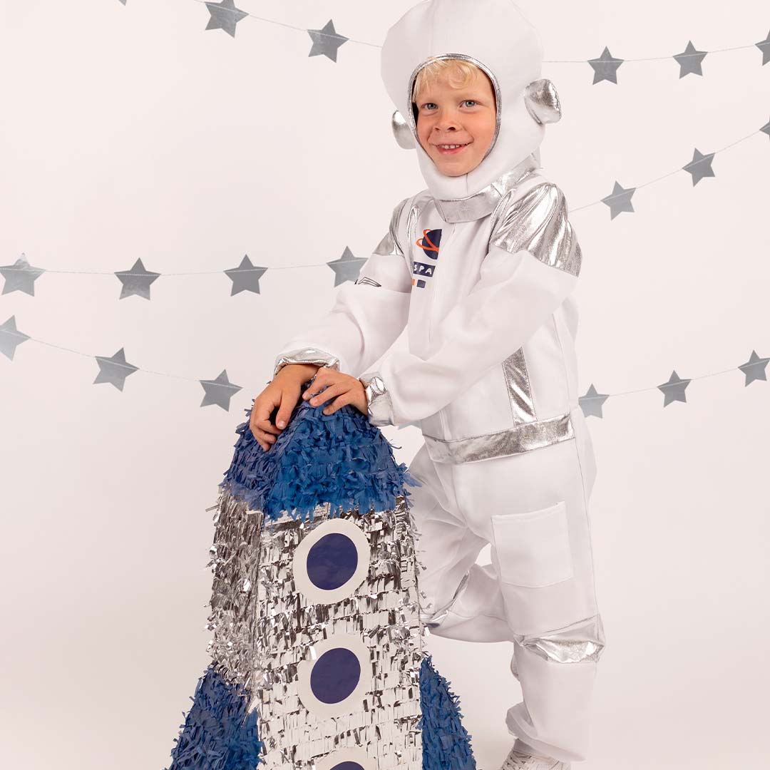 Partydeco astronaut verkleedkleding gedragen door een kind dat leunt op een blauw-zilveren raket piñata.