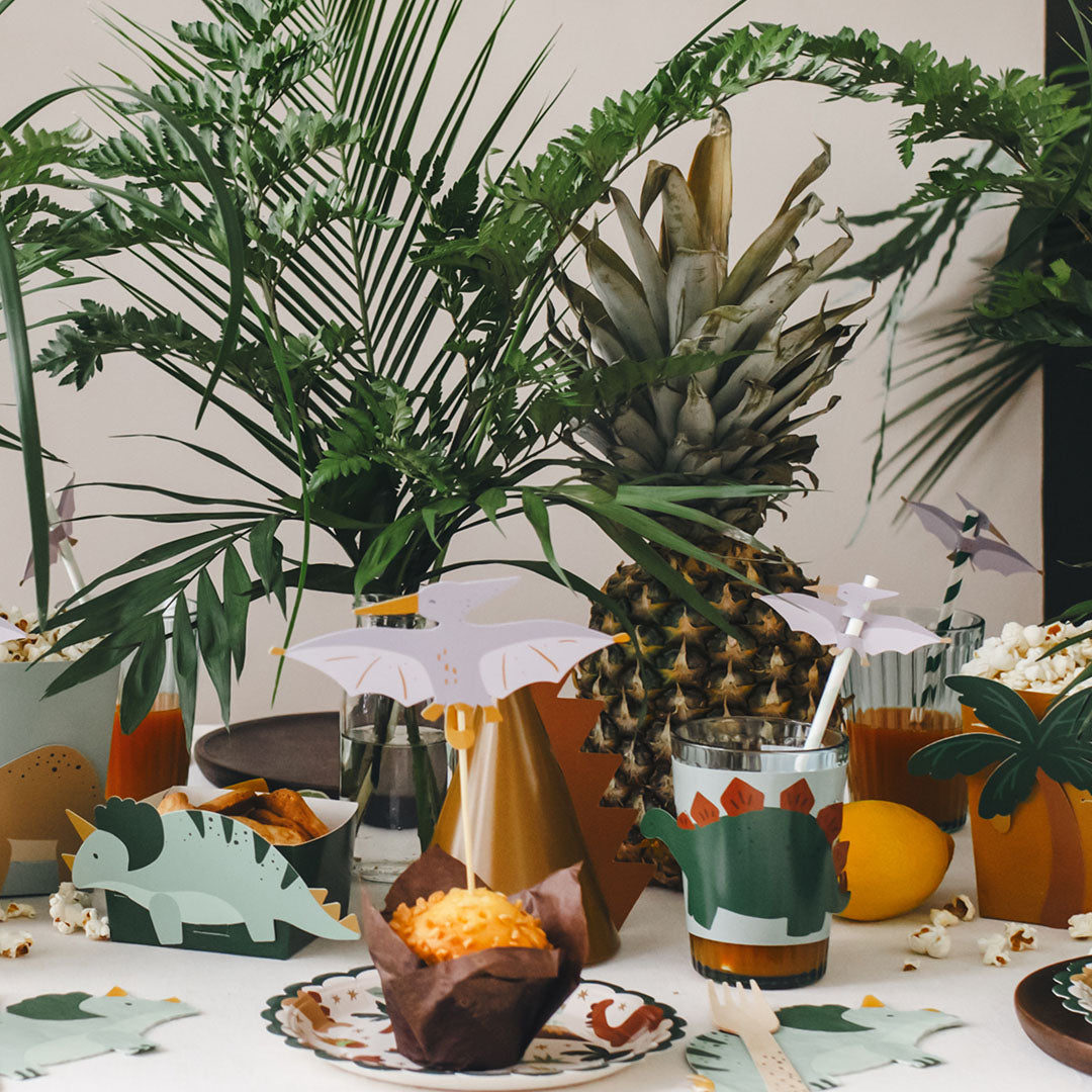 Een feestelijk gedekte tafel met partydeco dinosaurus feestartikelen, waaronder bekerhoesjes, bordjes en rietjes.