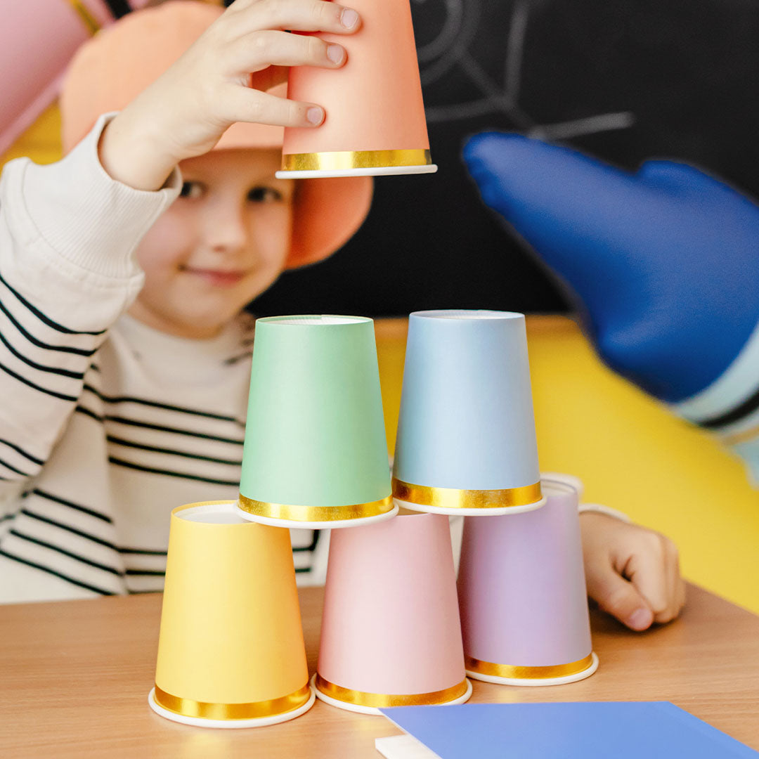 Een kind stapelt partydeco pastelkleurige papieren bekers met gouden rand tot een piramide.
