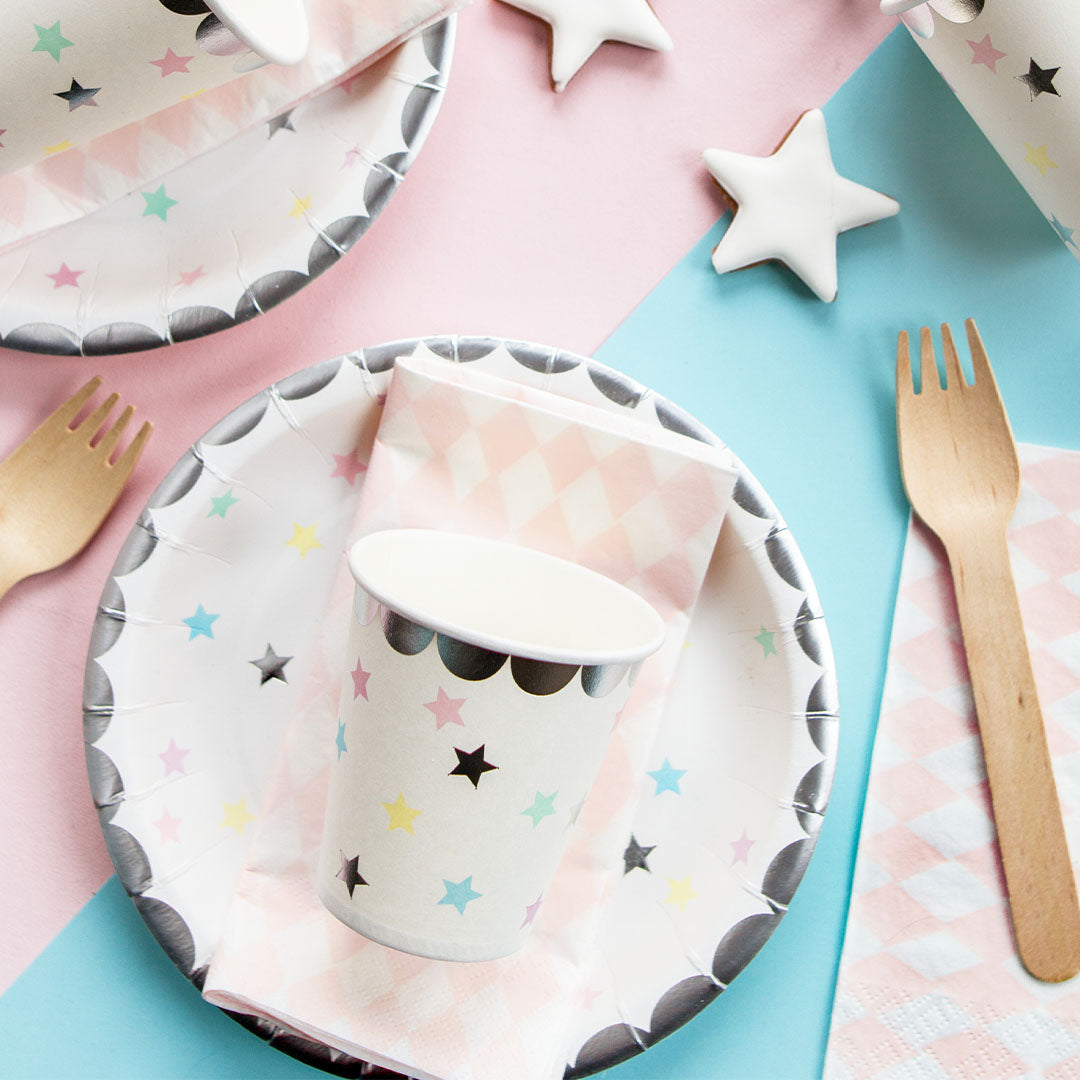 Partydeco papieren beker en bord met sterrenpatroon, samen met een servet en houten vork op een roze en blauwe ondergrond.