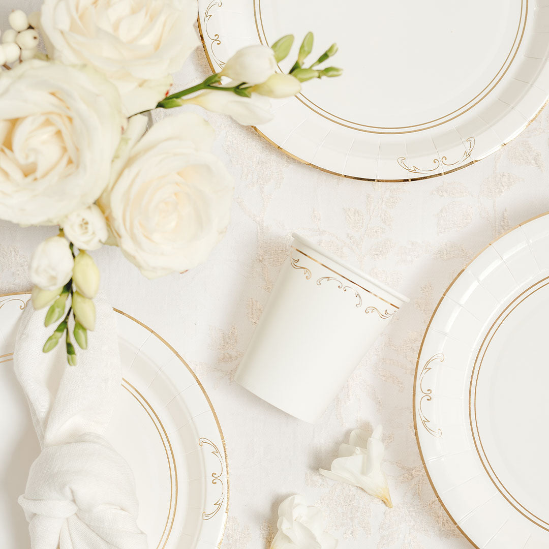Bovenaanzicht van een partydeco witte papieren partybeker met gouden ornamentpatroon, omringd door bijpassende borden en bloemen.