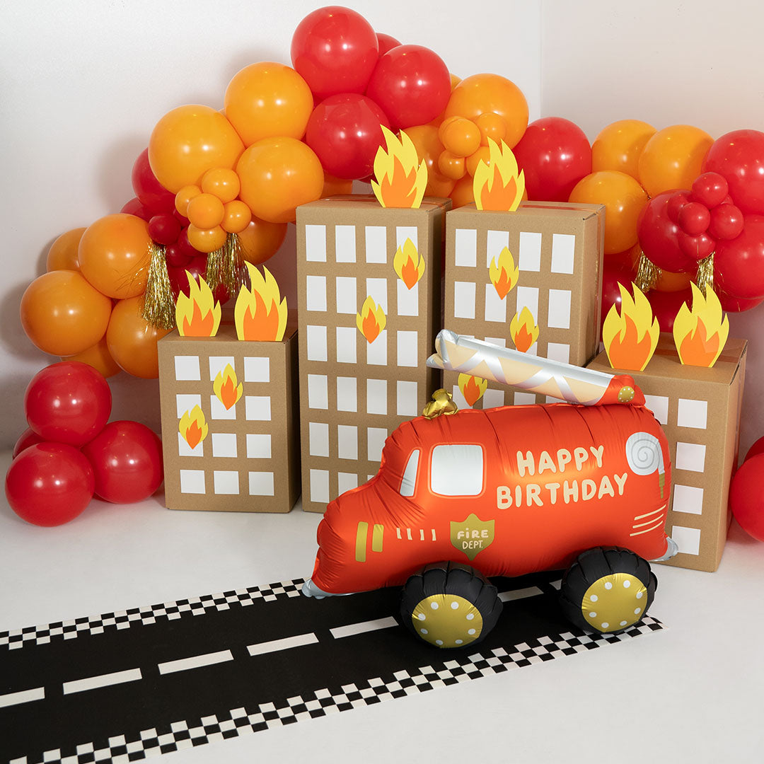 Partydeco folieballon brandweerwagen op een wegmat, omringd door ballonnen en kartonnen gebouwen als feestdecoratie.