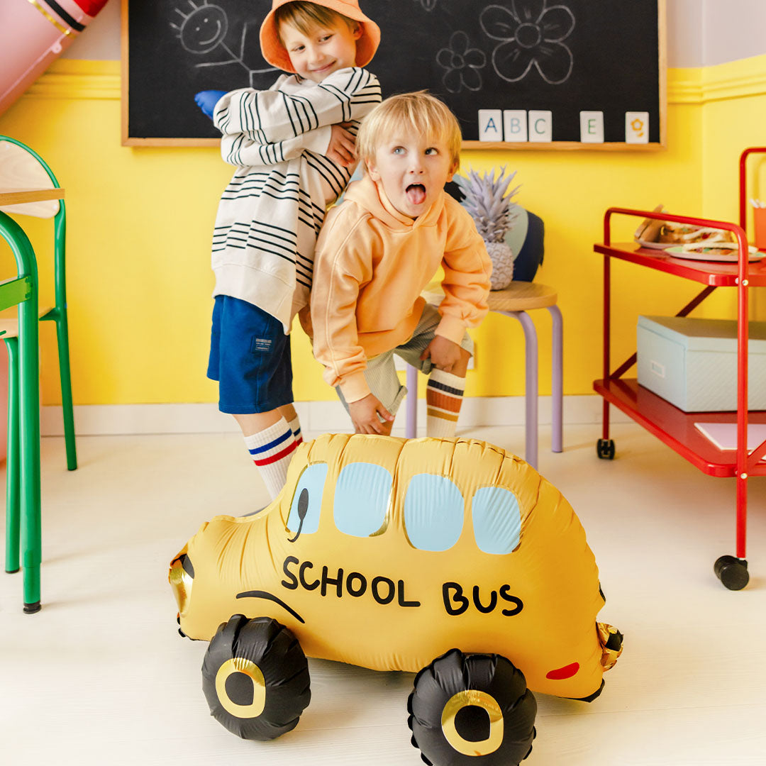 Twee kinderen poseren met de Partydeco folieballon in de vorm van een gele schoolbus in een klaslokaal.
