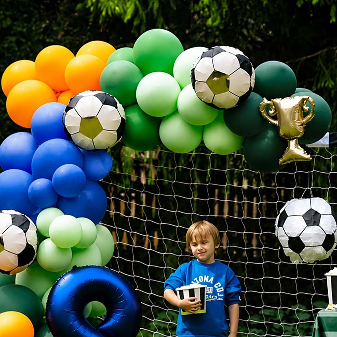 Jongen bij een feestelijke ballonnenboog met PartyDeco voetbalballonnen en een gouden trofee folieballon, voor een voetbaldoel.
