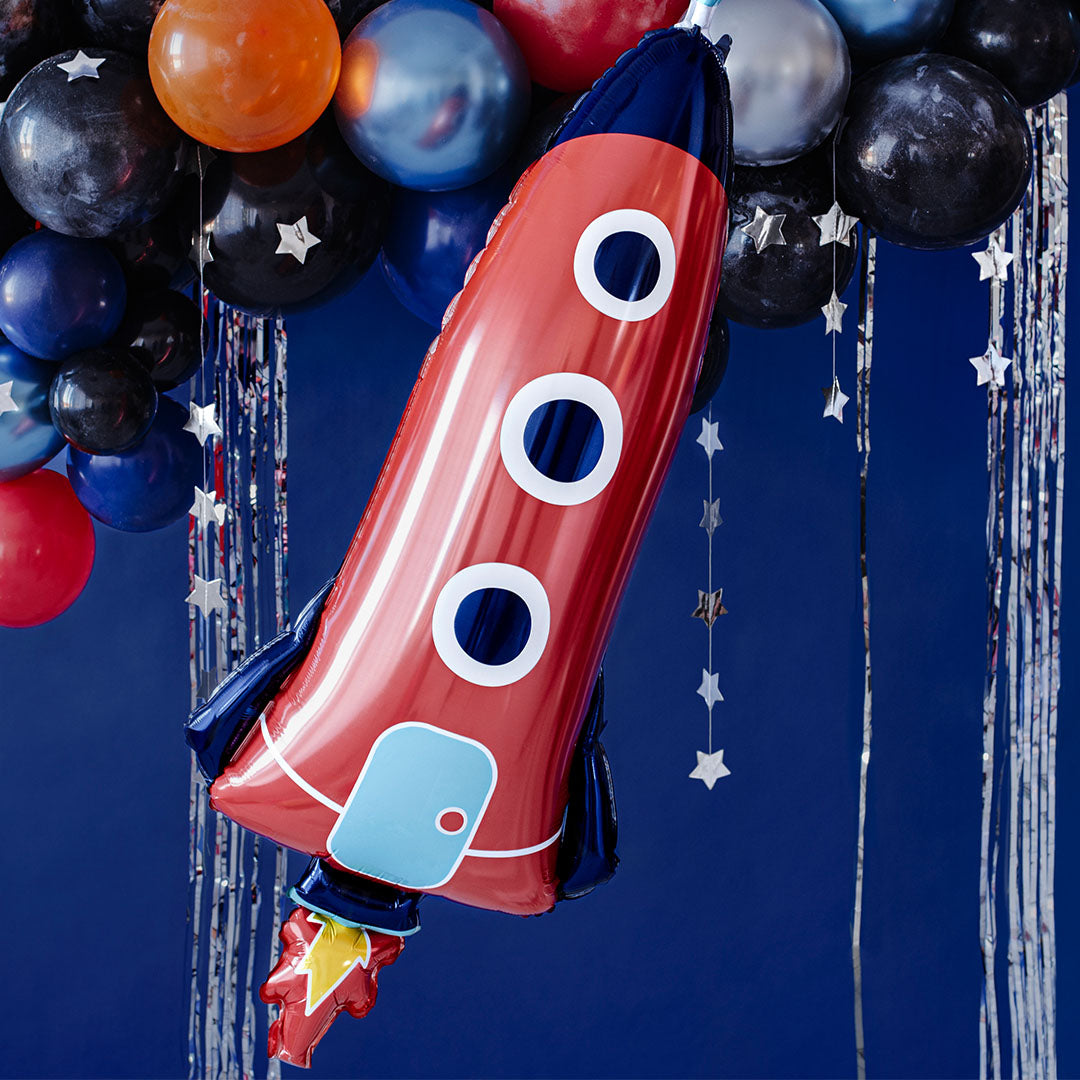 Partydeco folieballon in de vorm van een rode raket hangend tussen andere ballonnen en slingers op een feestje.