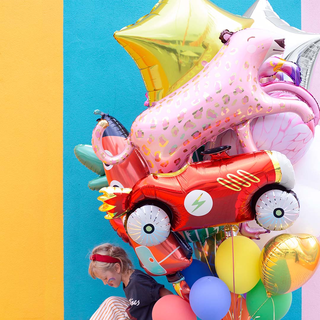 Kind zit naast een verzameling feestballonnen, waaronder een partydeco gouden ster folieballon, een rode auto en een roze luipaard.