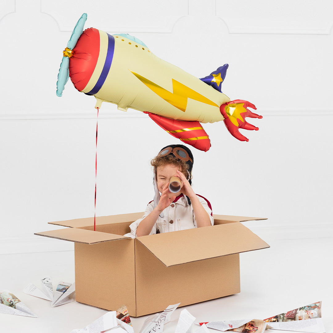 Een kind in een kartonnen doos speelt piloot onder de partydeco folieballon in de vorm van een vliegtuig.