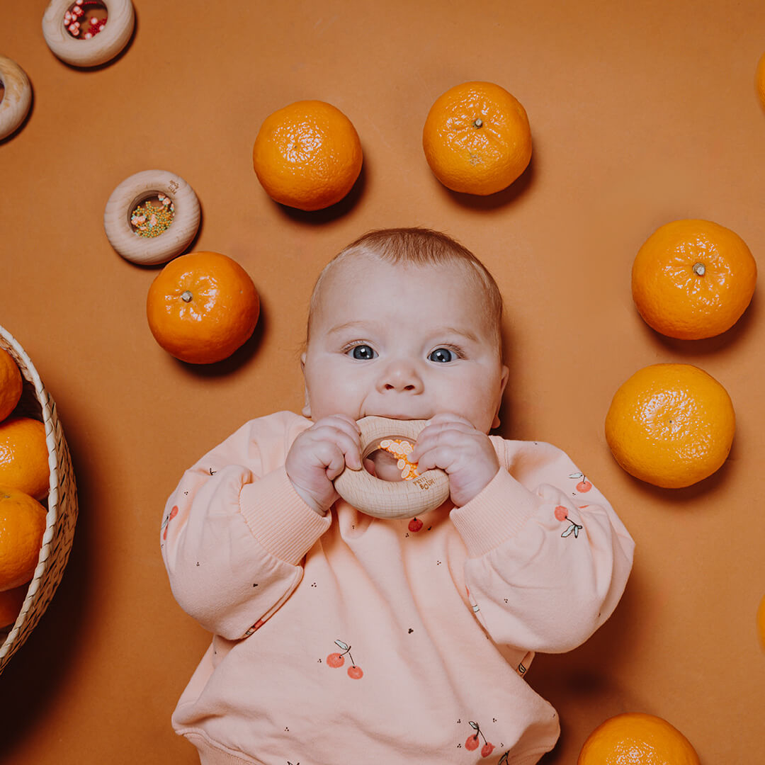 Een baby ligt op een bruine ondergrond en bijt op een Petit Boum houten bijtring, omringd door mandarijnen.