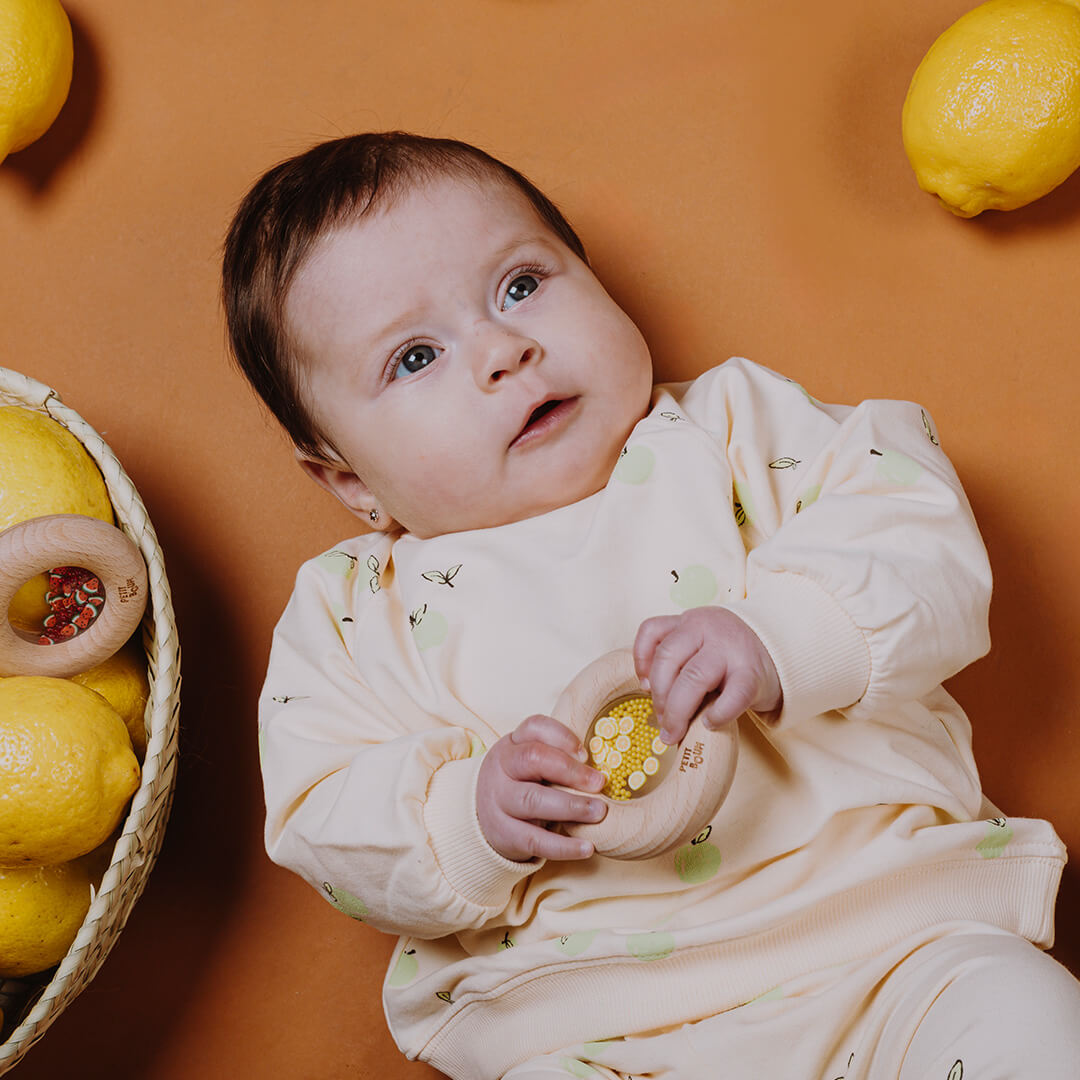 Een baby houdt een Petit Boum houten rammelaar met citroenen vast, liggend tussen echte citroenen.