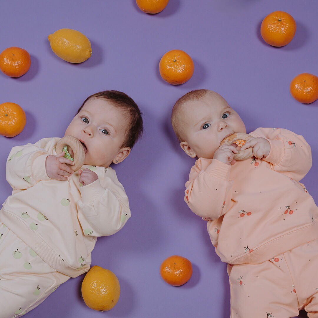 Twee baby's liggen op een paarse ondergrond en bijten op houten rammelaars tussen fruit.