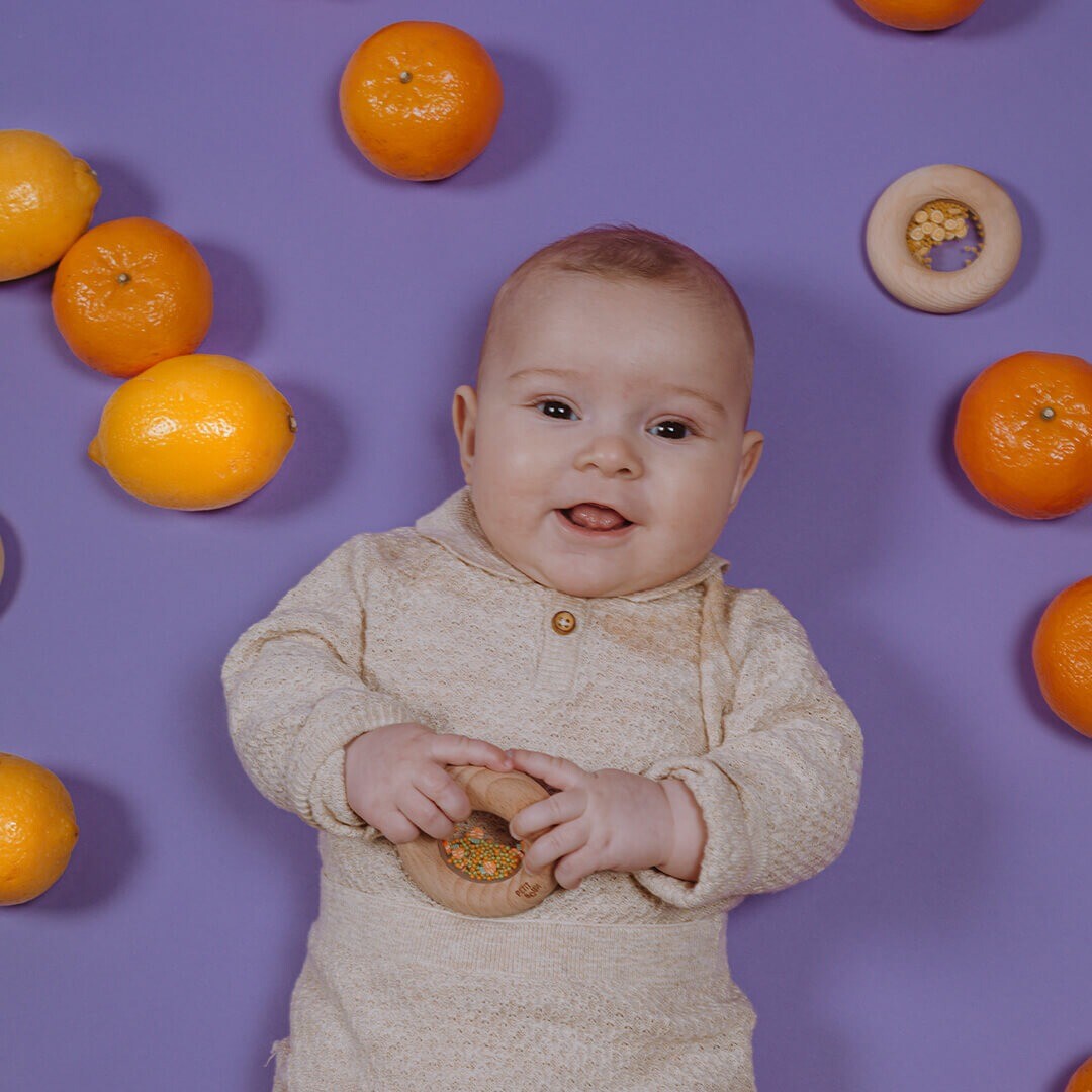 Een lachende baby ligt op een paarse ondergrond tussen fruit en houdt een houten bijtring vast.