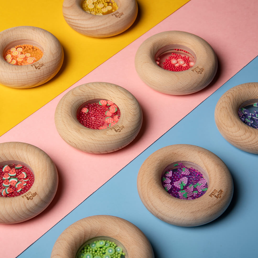 Meerdere Petit Boum houten bijtringen en rammelaars met verschillende fruitvullingen liggen op een kleurrijke ondergrond.