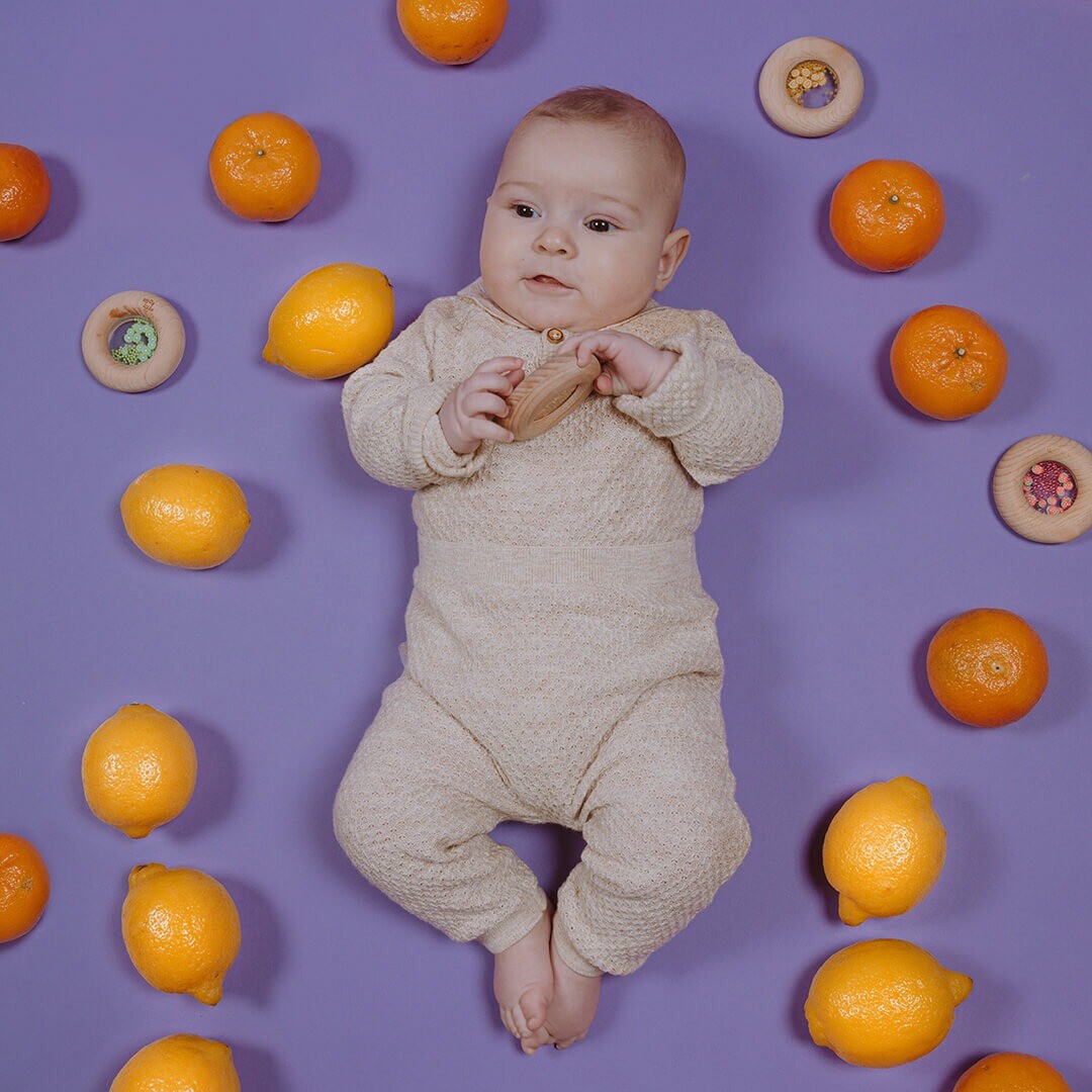 Een baby ligt op een paarse ondergrond en houdt een houten bijtring vast, omringd door citroenen, mandarijnen en andere bijtringen.