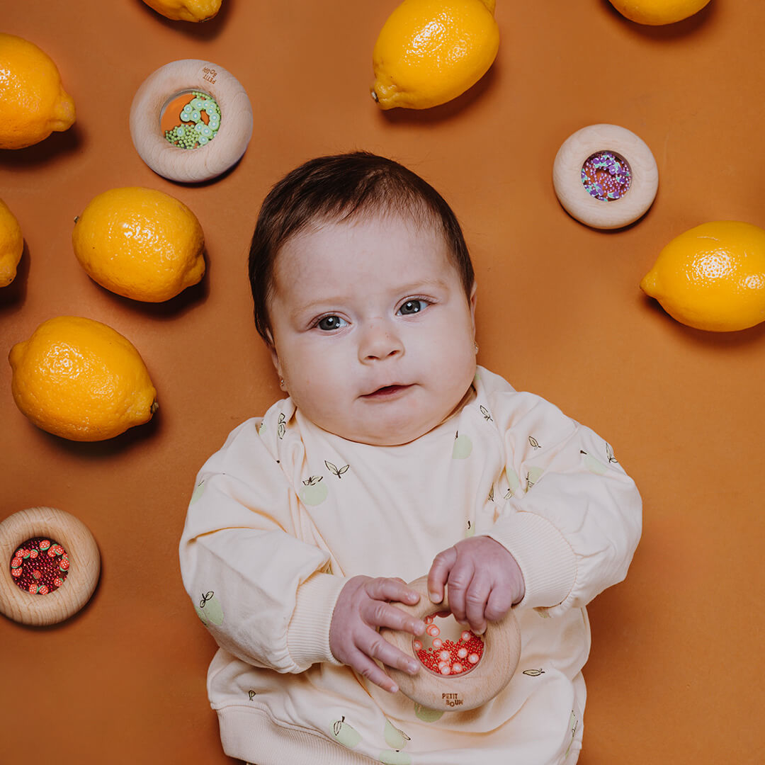 Een baby ligt op een bruine ondergrond en houdt een Petit Boum houten bijtring en rammelaar met rode tomaten vast, omringd door citroenen en andere rammelaars.