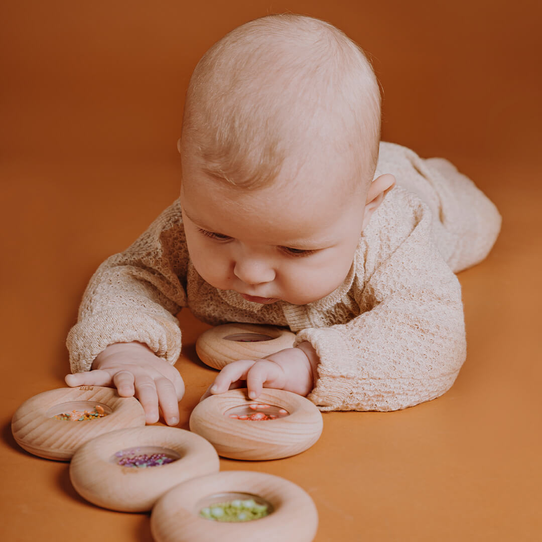 Een baby ligt op de buik en speelt met meerdere Petit Boum houten bijtringen en rammelaars met verschillende vullingen.