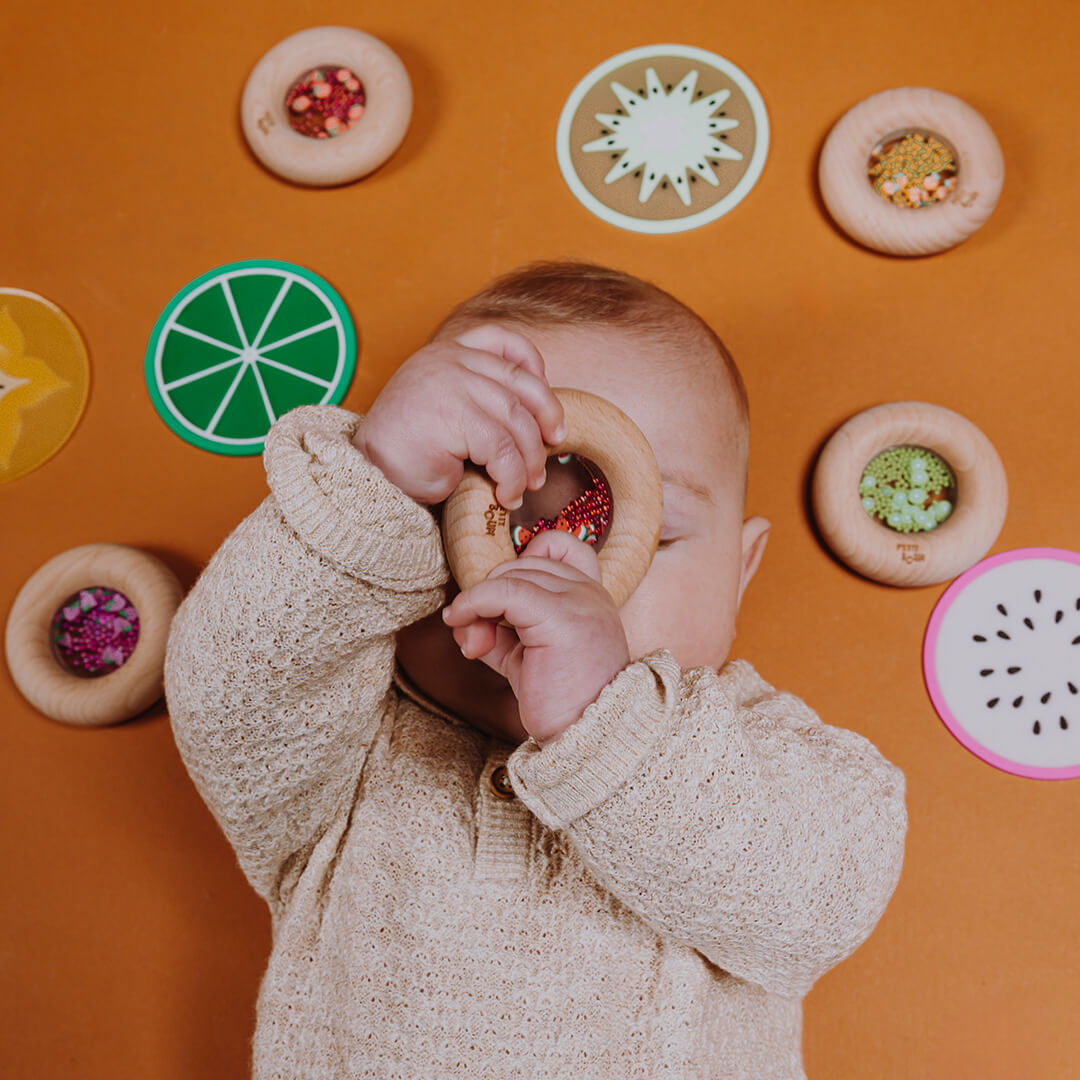 Een baby speelt met een Petit Boum houten bijtring en rammelaar, omringd door andere fruit-thema babyspeeltjes op een bruine ondergrond.
