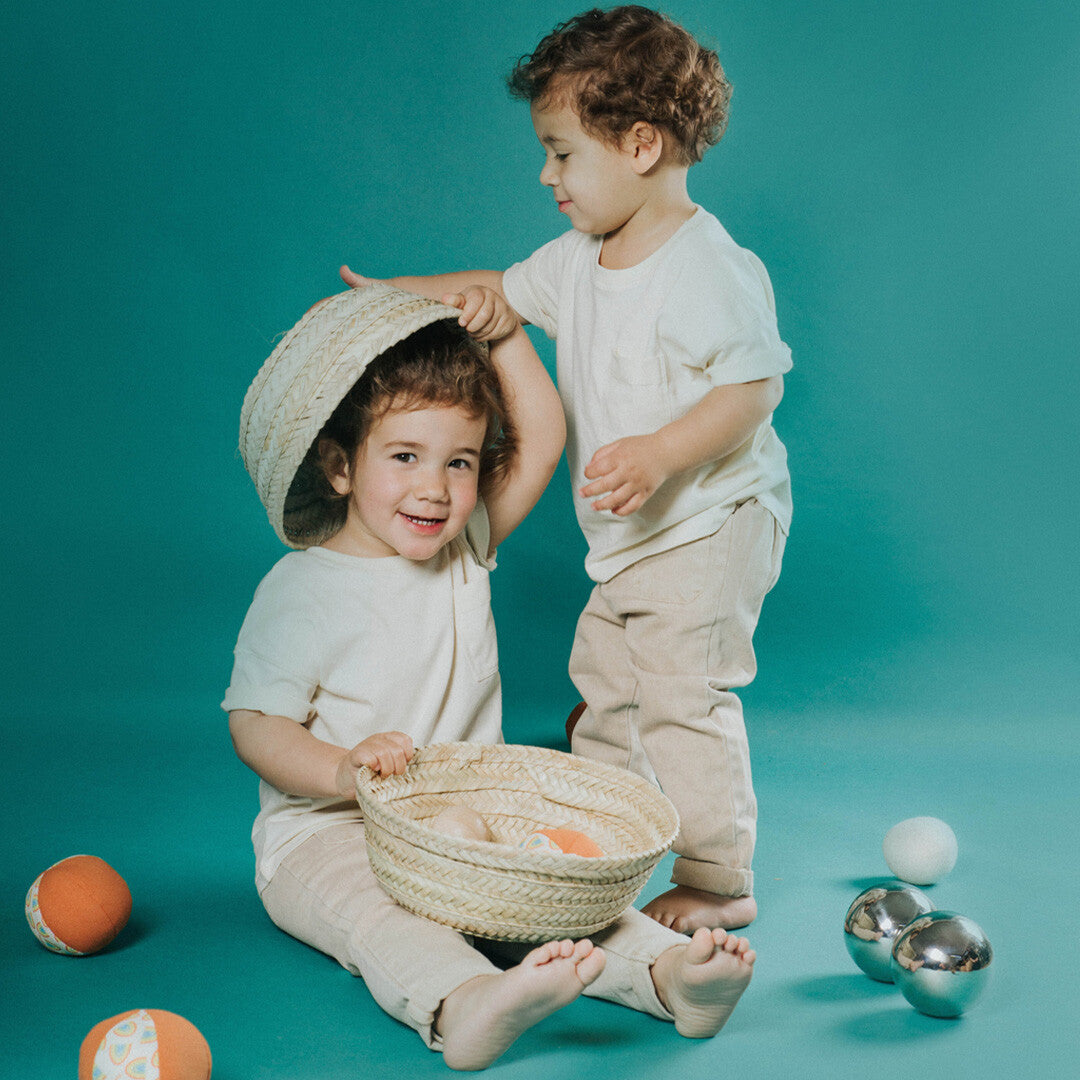 Twee kinderen spelen met de palmblad mand en sensorische ballen op een teal achtergrond.