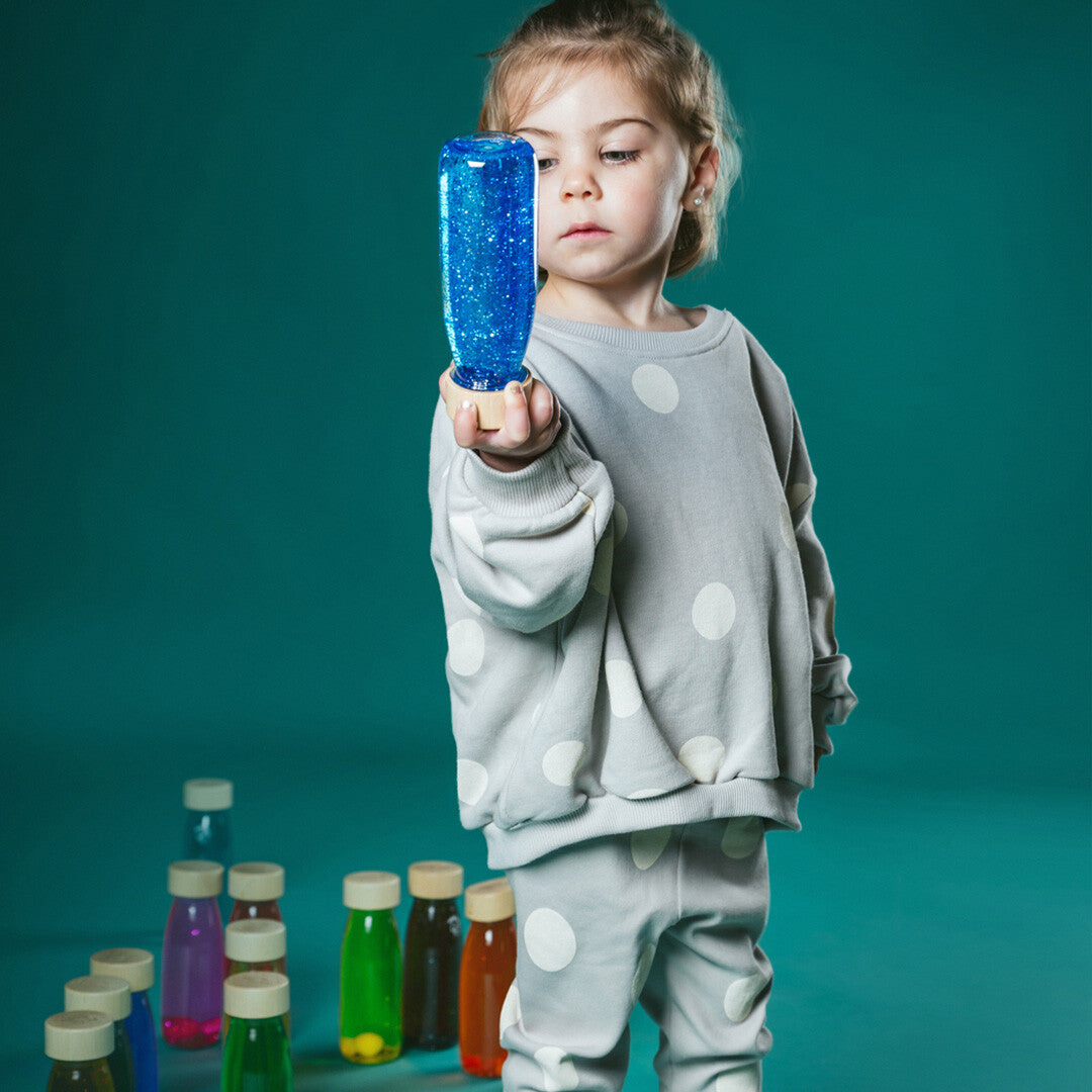 Een kind houdt een Petit Boum blauwe sensorische fles vast en kijkt ernaar, met andere gekleurde flessen op de achtergrond.