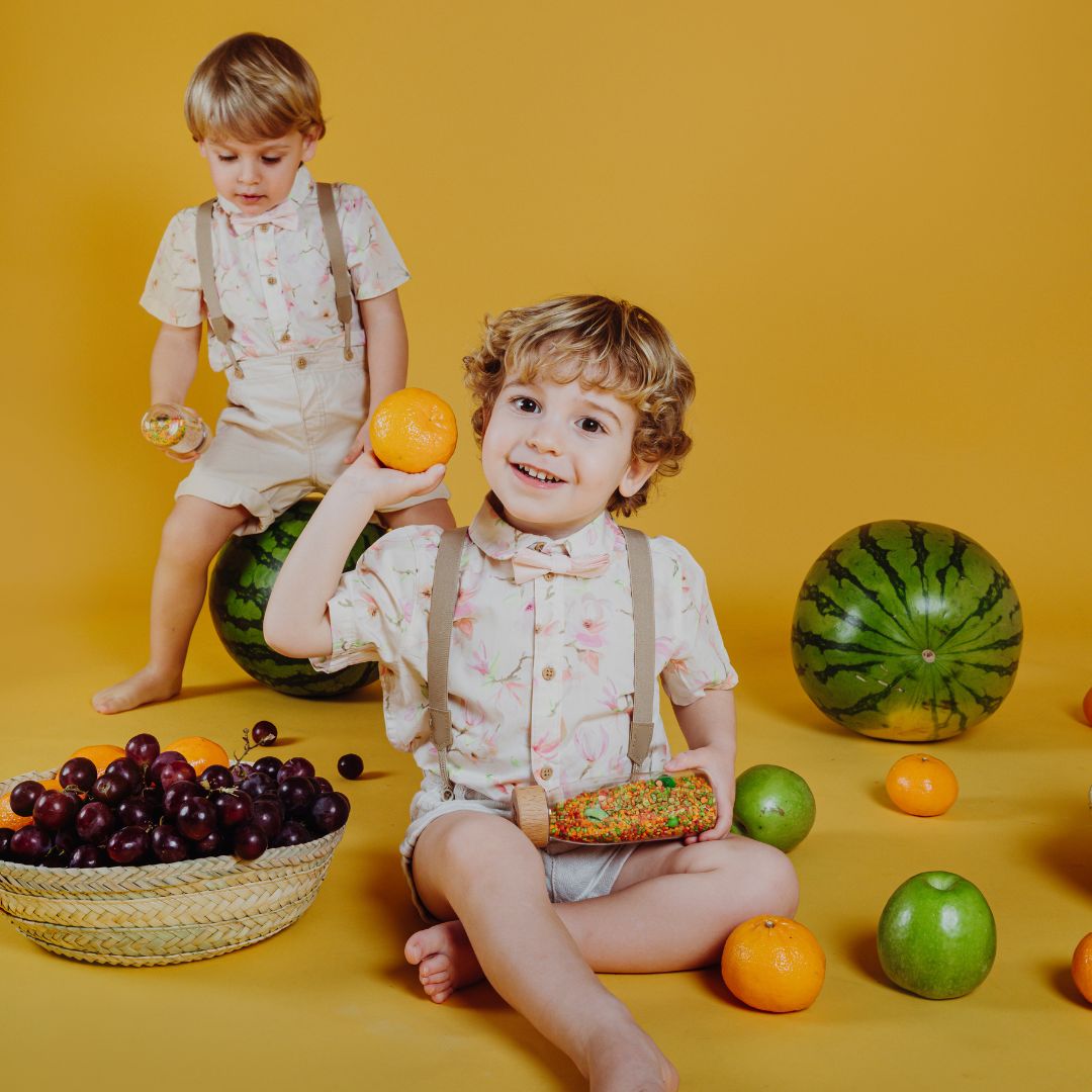Twee lachende jongens spelen met fruit en een Petit Boum sensorische fles op een gele achtergrond.