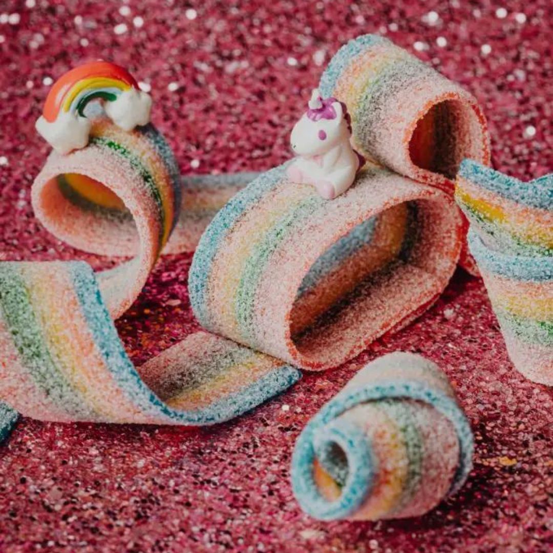 Regenboogkleurige snoepstrips met suiker en kleine eenhoorn- en regenboogfiguurtjes op een roze glitterachtergrond.
