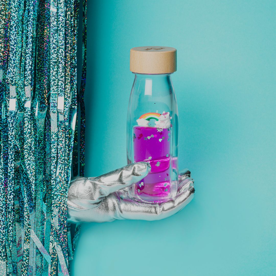 Een hand met zilveren handschoen houdt de petit boum sensorische fles met eenhoorn en regenboog vast, voor een blauwe glitterachtergrond.