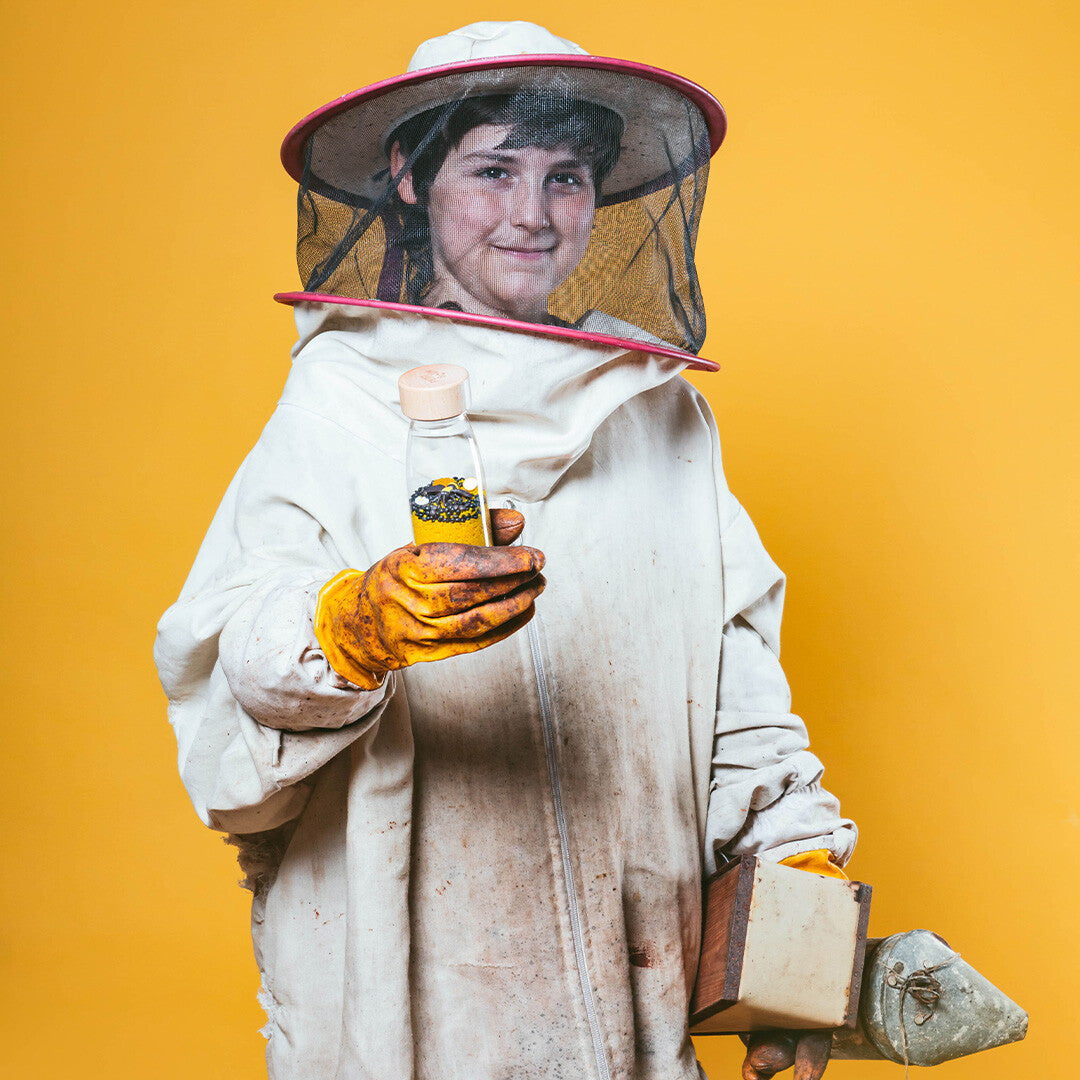 Een lachend kind in een bijenpak toont de Petit Boum sensorische fles met bijen en een bijenrookblazer.