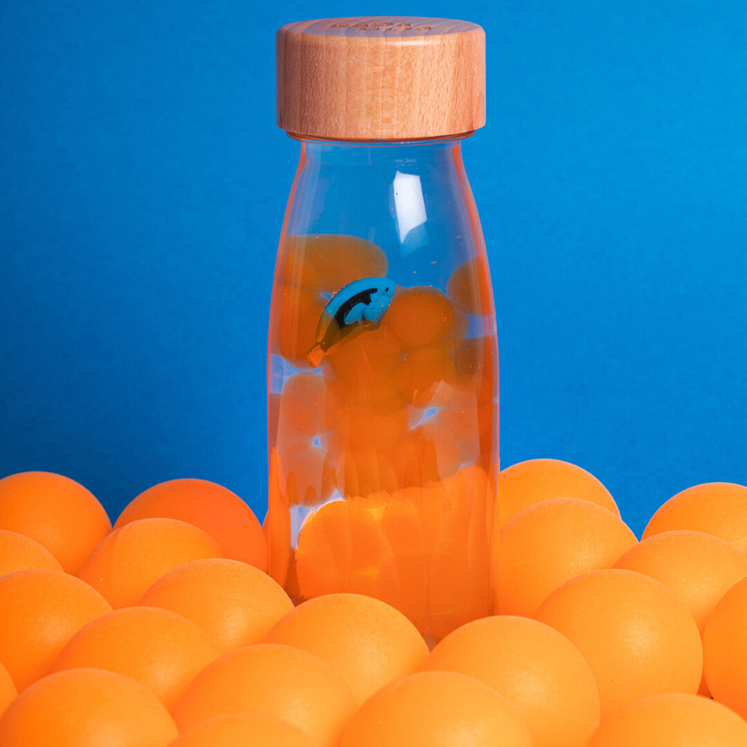 Petit Boum sensorische fles met blauwe tang vis en oranje balletjes, staand tussen meer oranje balletjes op een blauwe achtergrond.