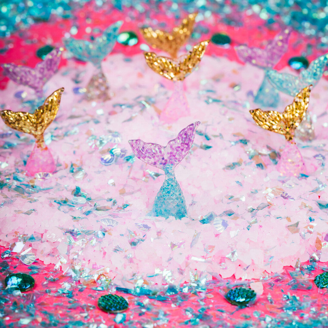 Detail van diverse zeemeerminstaartjes in goud, paars en blauw, omringd door roze en iriserende glitters.