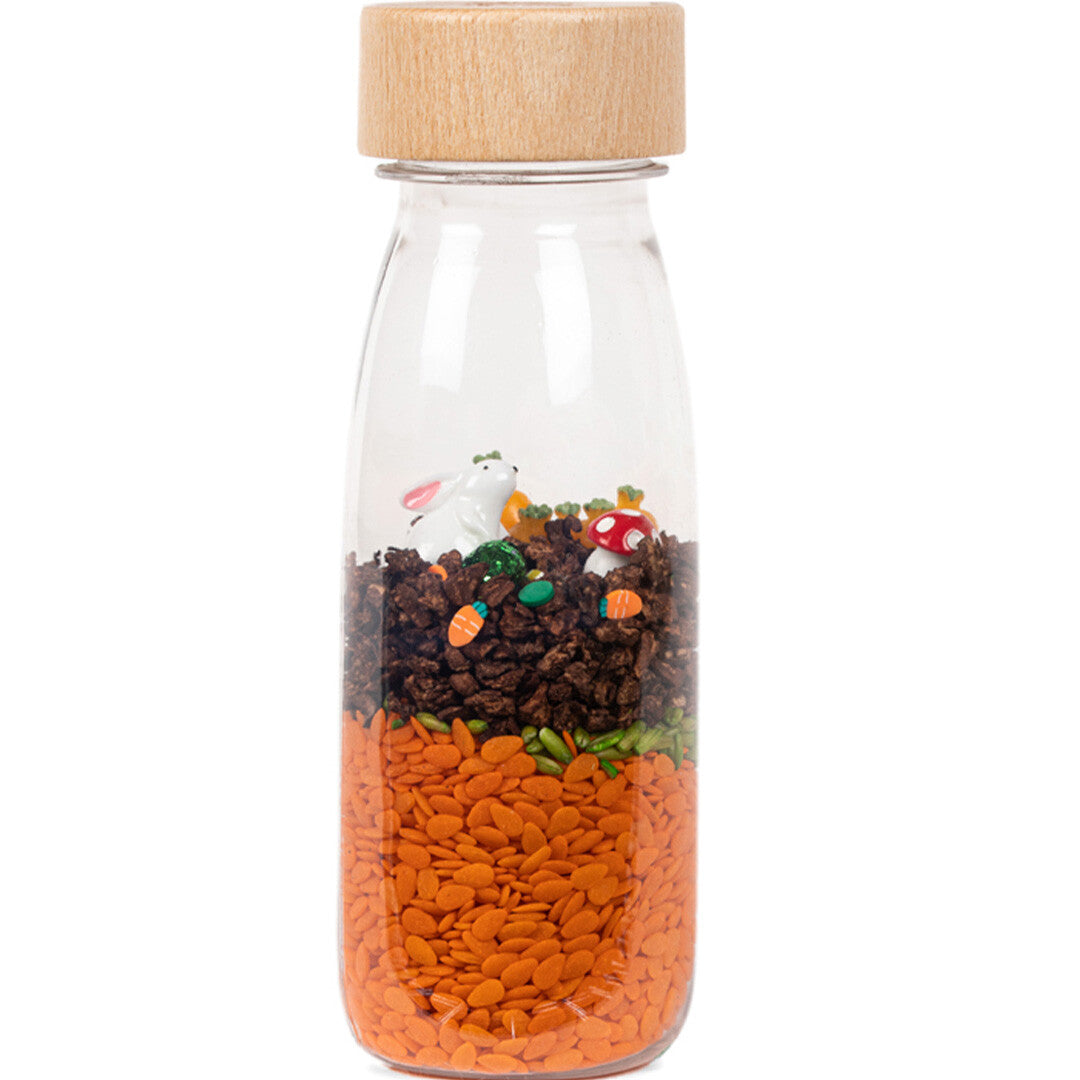 Petit Boum sensorische fles met een konijn, wortels en paddenstoelen op een ondergrond van oranje en bruine korrels.