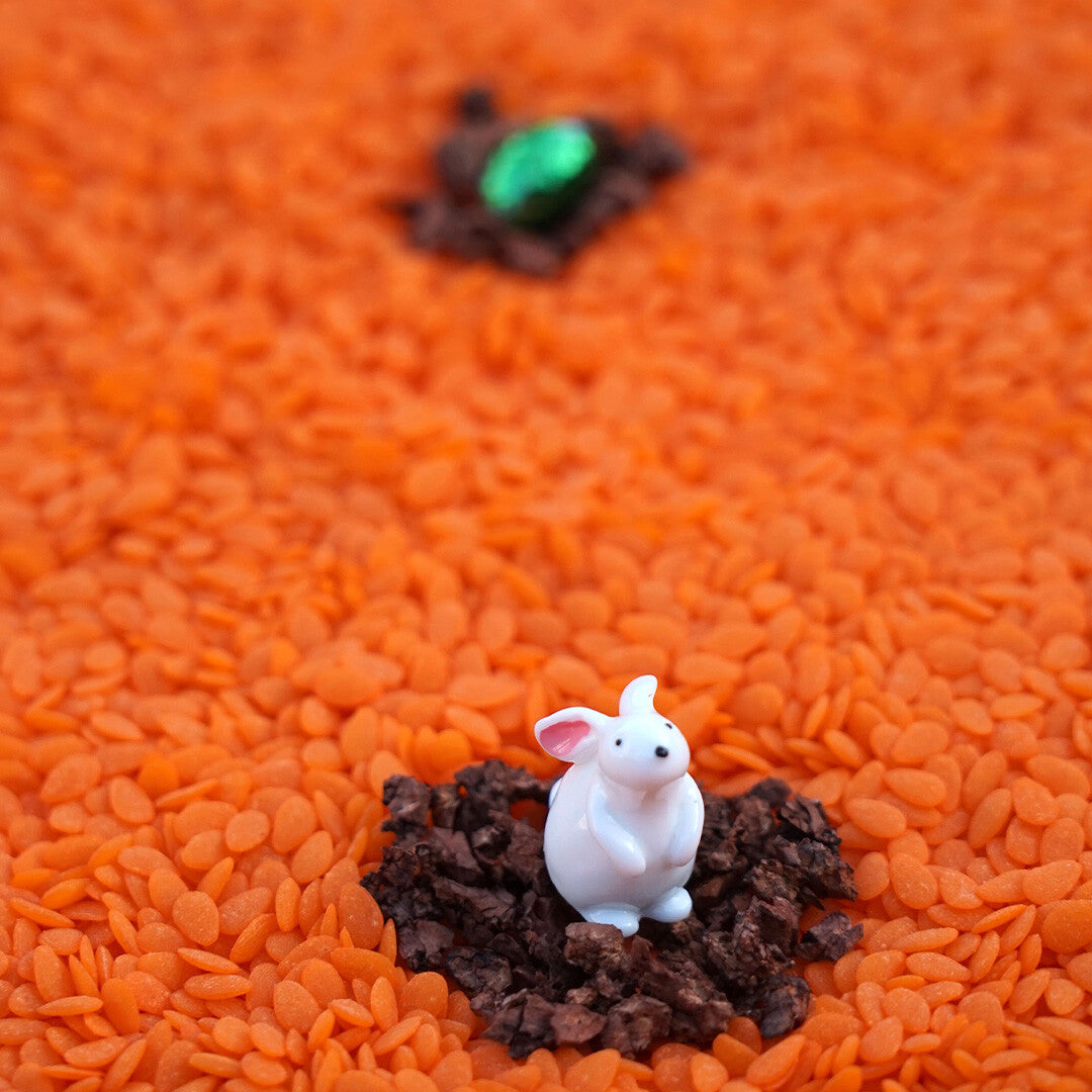 Detail van een wit konijnfiguurtje op bruine aarde, omringd door oranje korrels.