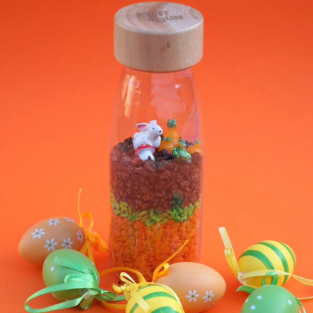 Petit Boum sensorische fles met konijn en wortels, omringd door paaseieren op een oranje achtergrond.