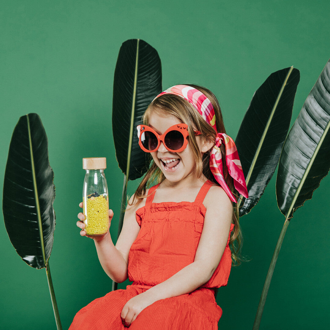 Een lachend meisje met zonnebril en hoofddoek houdt de petit boum sensorische fles vast voor een groene achtergrond.