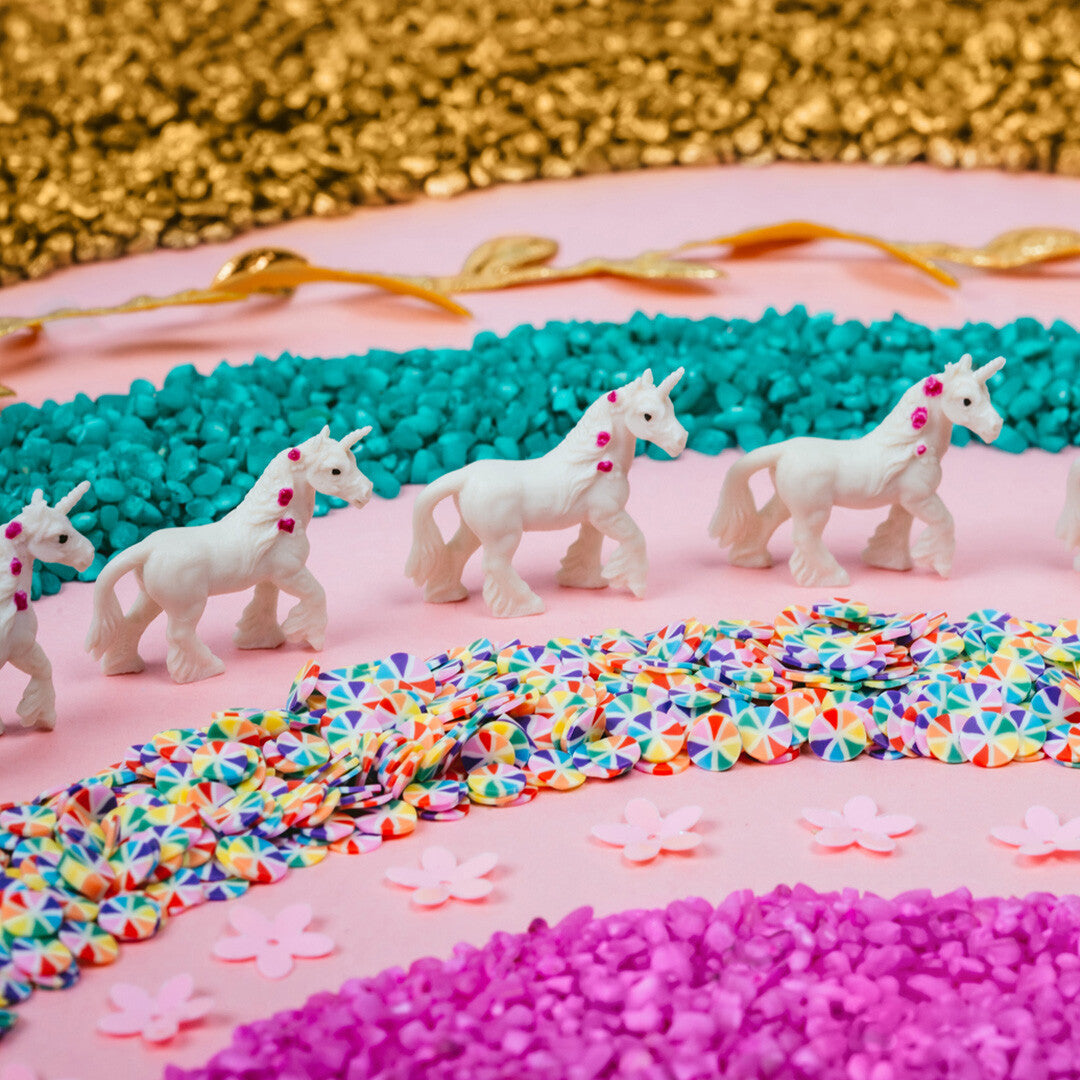 Witte eenhoornfiguren met roze hartjes, turquoise steentjes en kleurrijke confetti op een roze ondergrond.