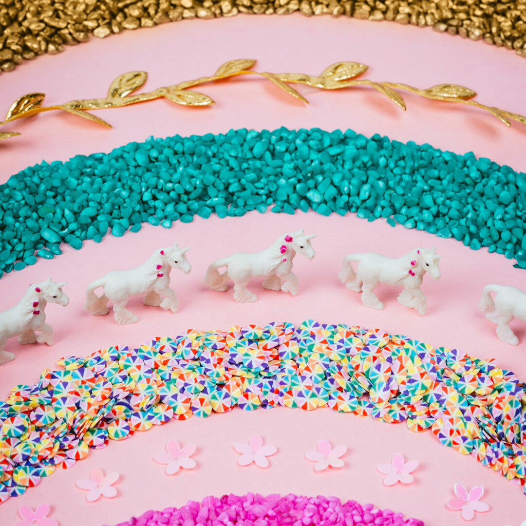 Witte eenhoornfiguren, turquoise steentjes, kleurrijke confetti en gouden bladeren op een roze ondergrond.
