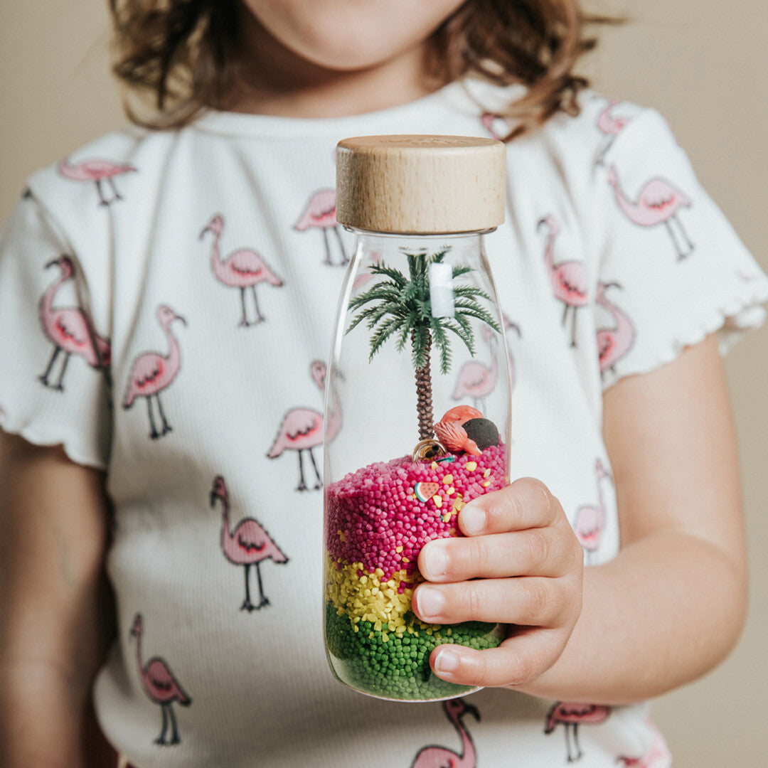 Een kinderhand houdt de petit boum sensorische fles met flamingo thema vast, met een flamingo-print shirt op de achtergrond.