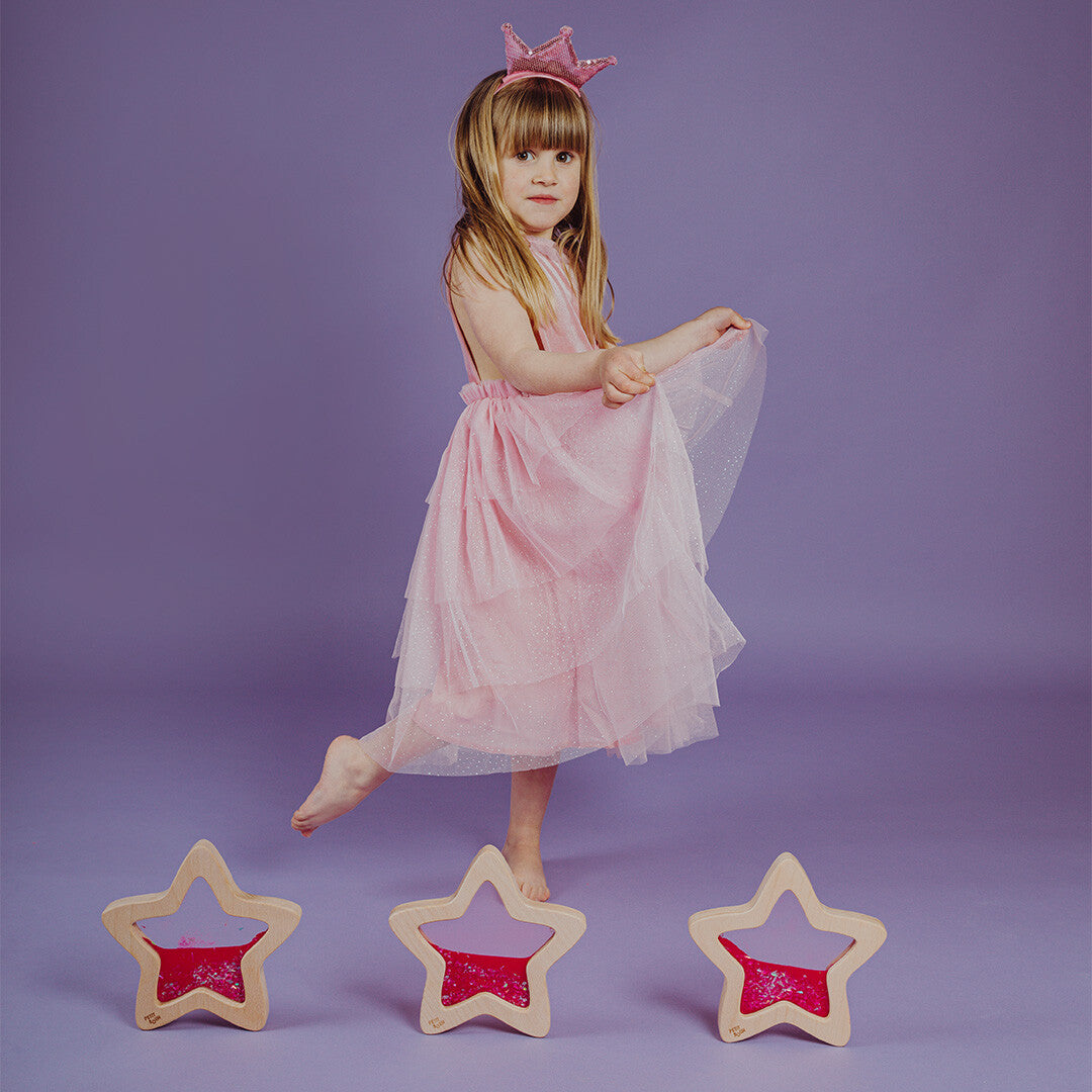 Een meisje in een roze jurk staat bij drie Petit Boum houten sensorische sterren.