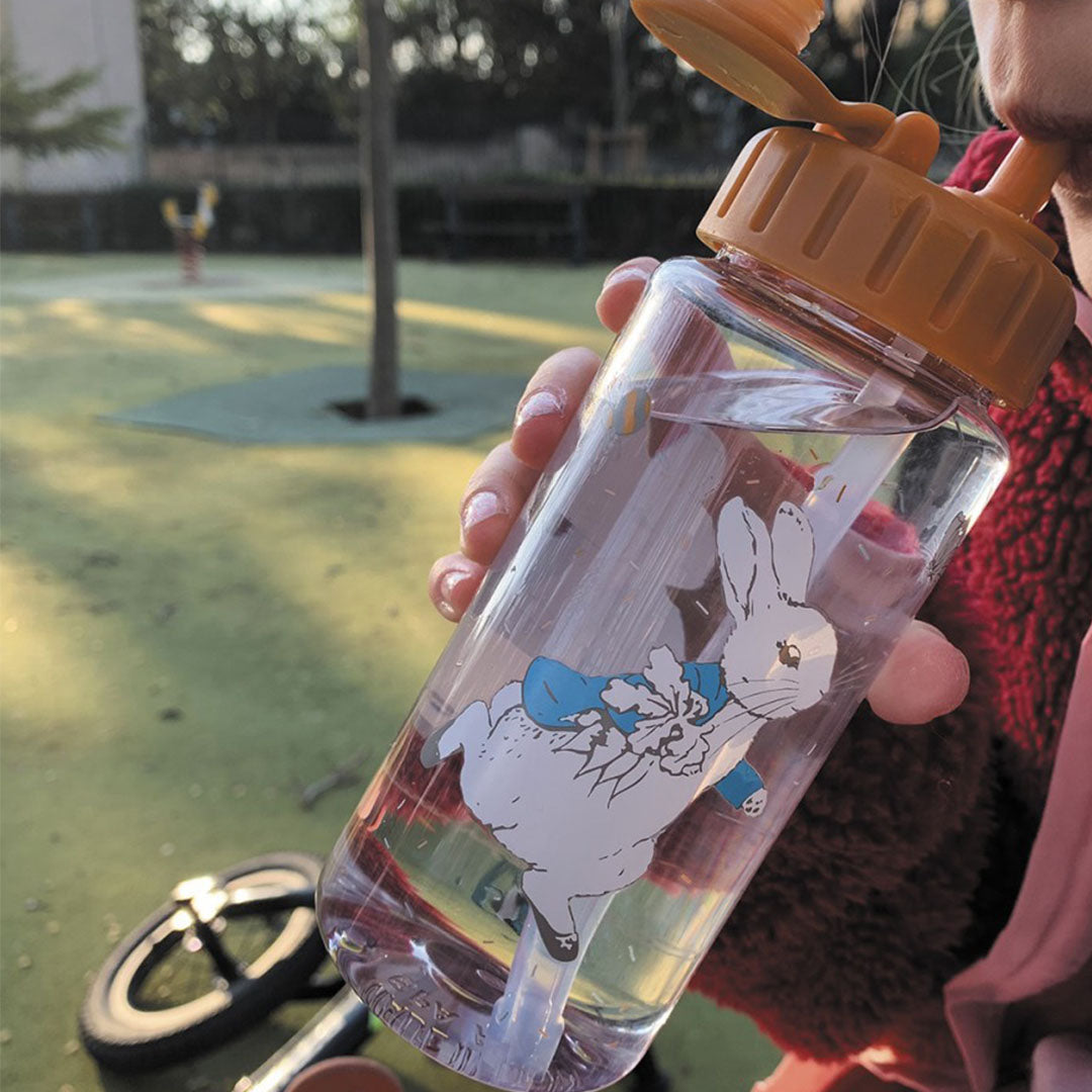 Een persoon drinkt uit de petit jour drinkfles met Pieter Konijn illustratie op een speelplaats.